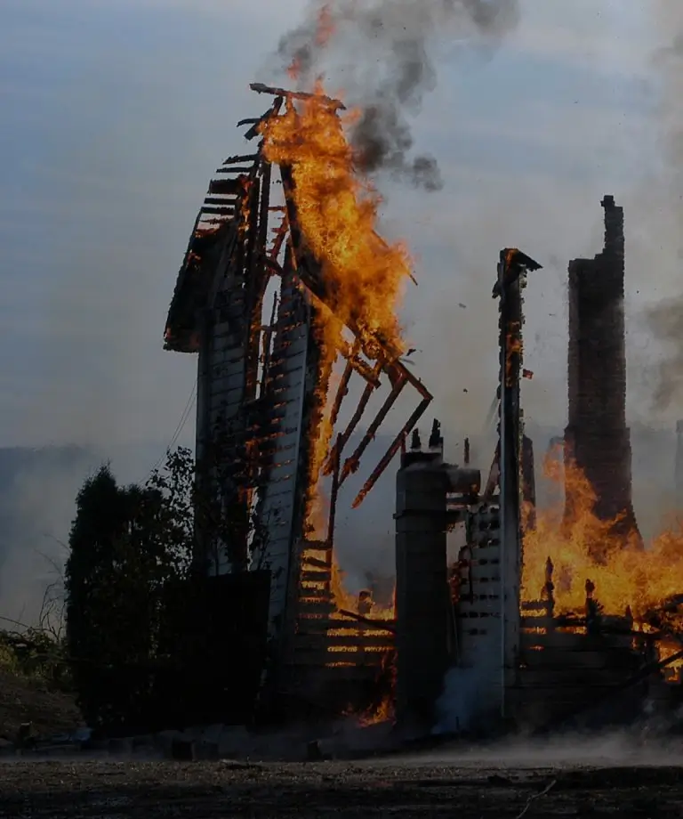 A large wooden structure engulfed in flames, emitting thick smoke against a dusk sky